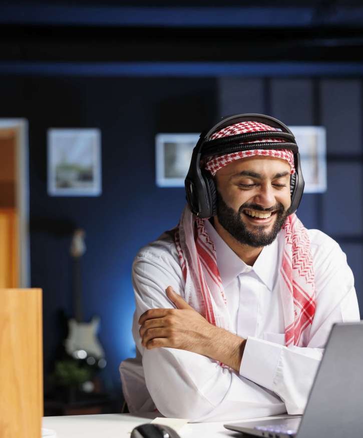 Utilizing wireless technology, Muslim man engages in online research and communication, showcasing his expertise and commitment to his profession. Arab guy using headphones and a laptop.